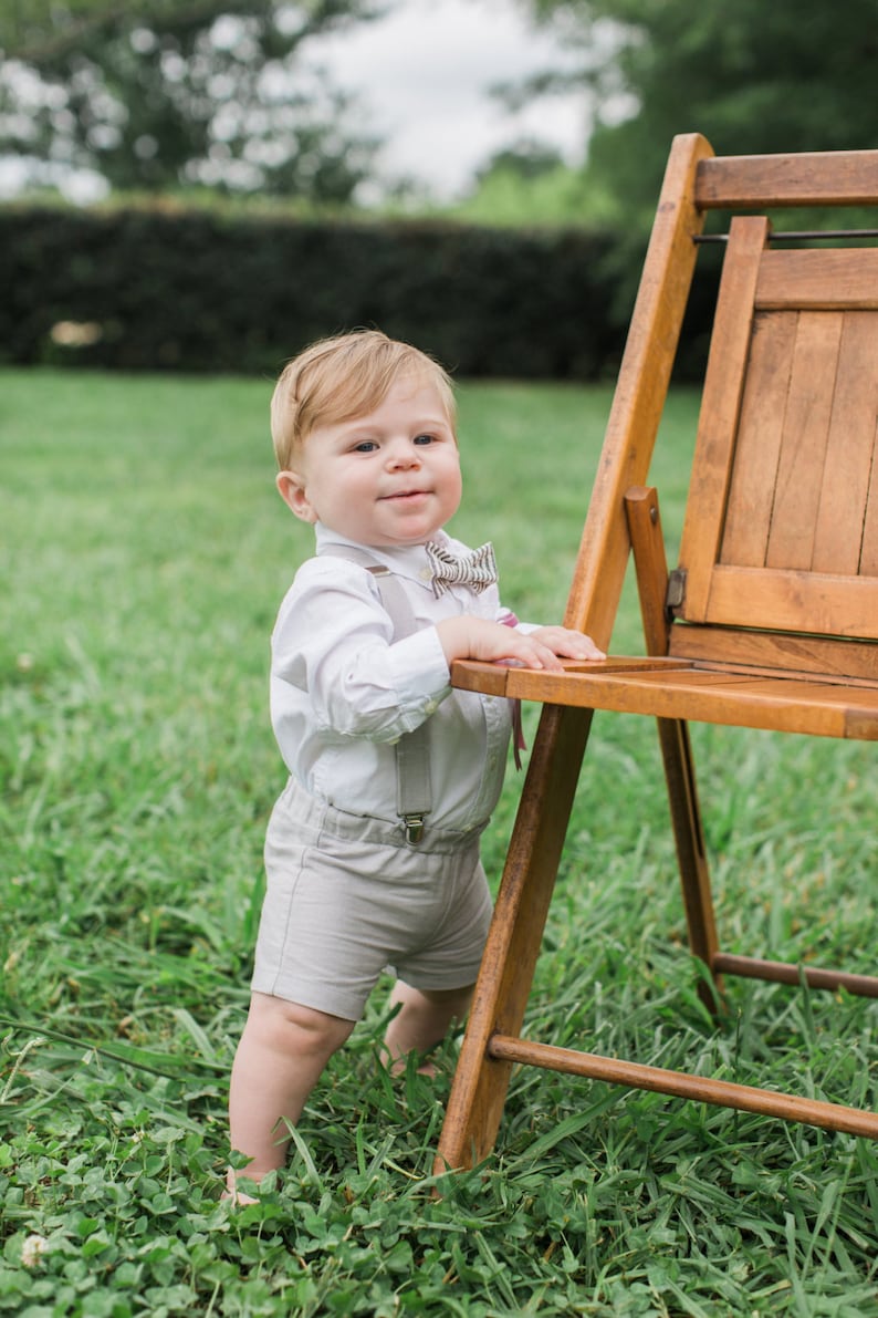 May include: A young child wearing a white button-down shirt, beige shorts, and a striped bow tie stands next to a wooden chair in a grassy field.