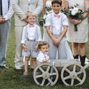 May include: Four young boys in matching white shirts and gray pants stand in front of a wooden wagon with white wheels. Two of the boys are wearing pink bow ties. The boys are all smiling and looking at the camera.