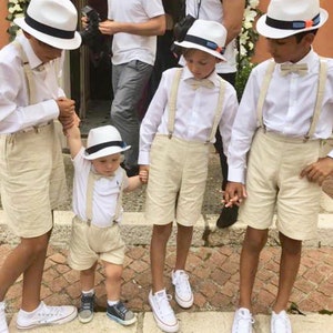 May include: Four young boys wearing white shirts, beige shorts, and white hats with black bands. They are all wearing suspenders and bow ties. The boys are standing on a brick walkway.