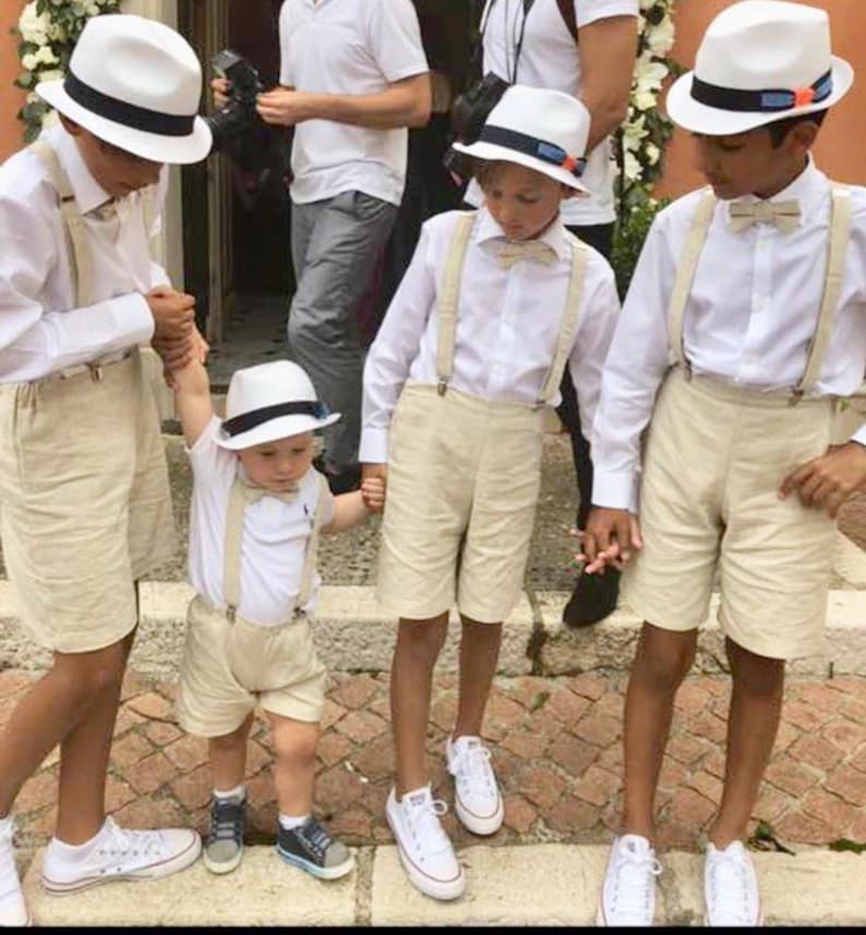 May include: Four young boys wearing white shirts, beige shorts, and white hats with black bands. They are all wearing suspenders and bow ties. The boys are standing on a brick walkway.