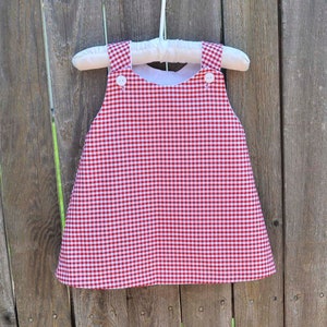 May include: A red and white gingham-patterned dress with white lining, hanging on a white padded hanger. The dress has two white buttons on each shoulder strap. The dress is a classic style, perfect for a child.