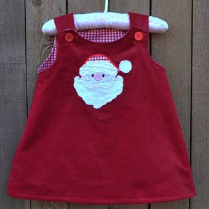 May include: Red corduroy dress with a white gingham lining and an embroidered Santa Claus face on the front.