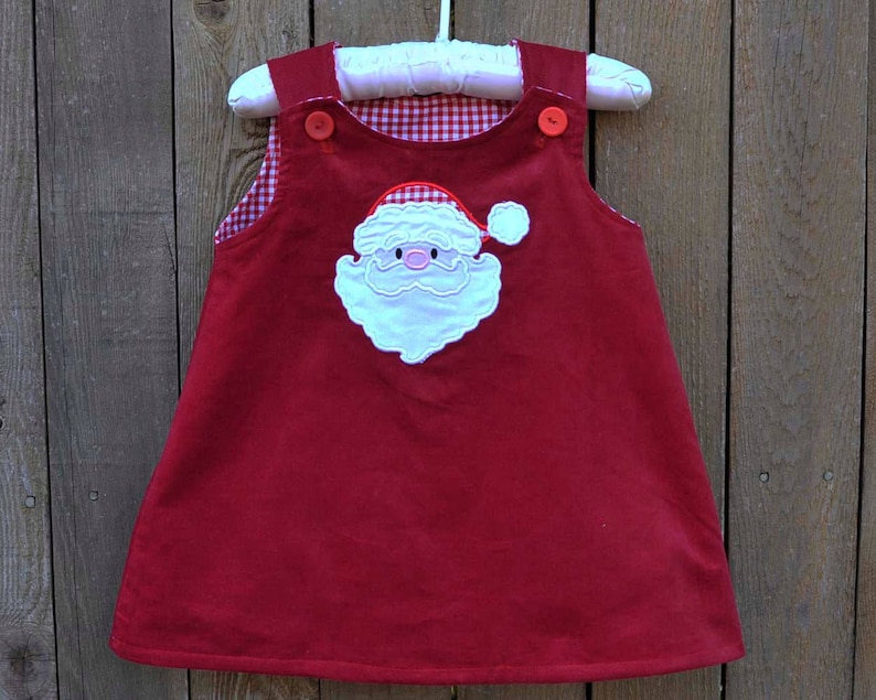 May include: Red corduroy dress with a white gingham lining and an embroidered Santa Claus face on the front.