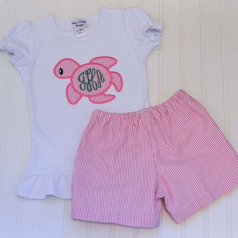 May include: A white short-sleeved shirt with a pink and green embroidered turtle with the letters "S.E.A." inside. The shirt is paired with pink and white striped shorts.