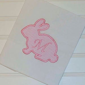 May include: A white, folded card featuring a pink embroidered bunny design. The bunny is filled with pink and white stripes and has a cursive letter "M" in the centre. The card is angled, revealing a portion of the white surface.