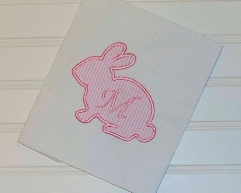May include: A white, folded card featuring a pink embroidered bunny design. The bunny is filled with pink and white stripes and has a cursive letter "M" in the centre. The card is angled, revealing a portion of the white surface.