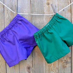 May include: Two pairs of shorts hanging on a clothesline. One pair is purple and the other is green.