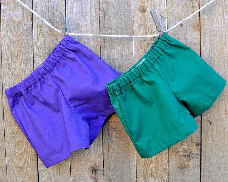 May include: Two pairs of shorts hanging on a clothesline. One pair is purple and the other is green.