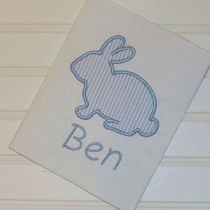 May include: White rectangular fabric featuring a light blue seersucker bunny applique. The bunny is outlined in blue thread, with the name "Ben" embroidered below in blue script. The fabric is set against a white background.