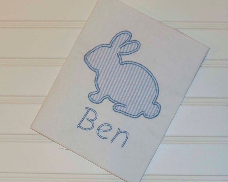 May include: White rectangular fabric featuring a light blue seersucker bunny applique. The bunny is outlined in blue thread, with the name "Ben" embroidered below in blue script. The fabric is set against a white background.