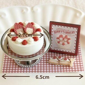 May include: A miniature strawberry shortcake with a white frosting base, topped with strawberries and cream swirls. The cake is 2.7 cm wide and sits on a silver cake stand. A sign that reads "NUUUFY'S BAKERY" is next to the cake. The base is 6.5 cm wide.