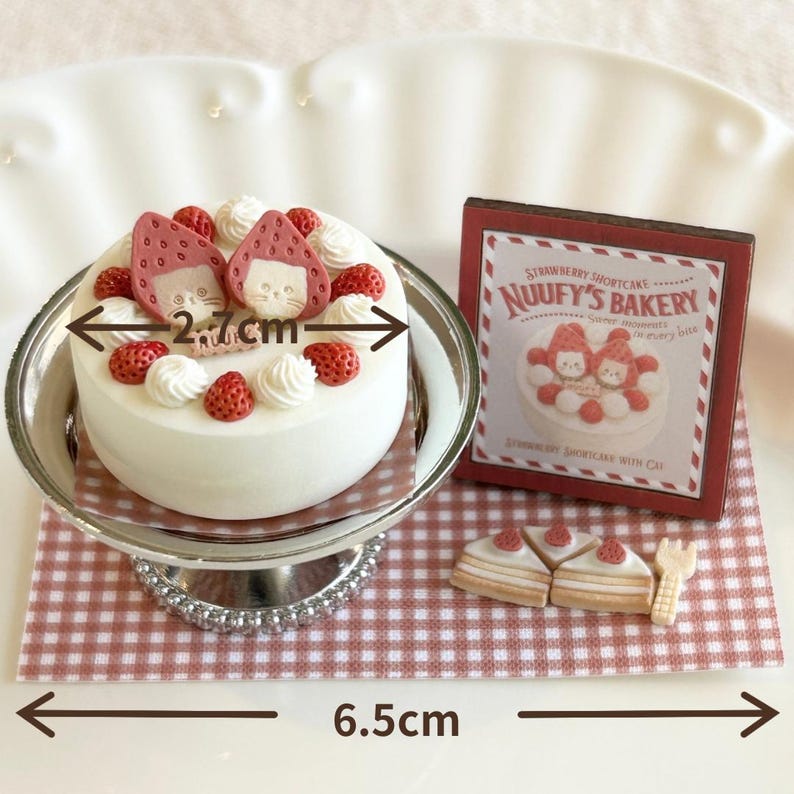 May include: A miniature strawberry shortcake with a white frosting base, topped with strawberries and cream swirls. The cake is 2.7 cm wide and sits on a silver cake stand. A sign that reads "NUUUFY'S BAKERY" is next to the cake. The base is 6.5 cm wide.