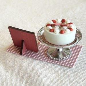 May include: A miniature white cake with whipped cream and red strawberry decorations sits on a silver cake stand. A small pink wooden frame rests on a checkered pink and white surface, suggesting a bakery or dessert theme.