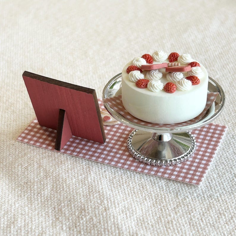 May include: A miniature white cake with whipped cream and red strawberry decorations sits on a silver cake stand. A small pink wooden frame rests on a checkered pink and white surface, suggesting a bakery or dessert theme.