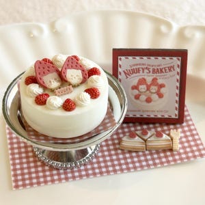 May include: A miniature strawberry shortcake with cat-shaped strawberry decorations, whipped cream, and fresh strawberries. The cake sits on a silver cake stand, next to a sign that reads "Nuuufy's Bakery". A miniature fork and cake slices are also present.