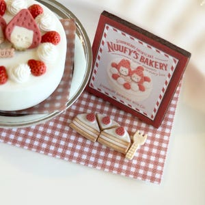 May include: A white cake decorated with strawberries and cream, alongside a box and cookies. The box features the text "NUUUFY'S BAKERY" and an illustration of a strawberry shortcake. The cookies are shaped like cake slices with strawberry toppings.