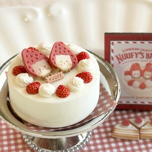 May include: A white cake decorated with strawberries and cream swirls, topped with two strawberry-shaped figures with cat faces. The cake sits on a silver cake stand, with a sign that reads "NUUFY'S BAKERY".