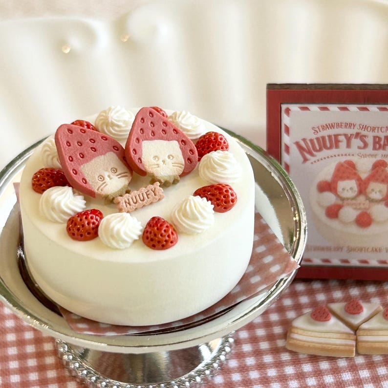 May include: A white cake decorated with strawberries and cream swirls, topped with two strawberry-shaped figures with cat faces. The cake sits on a silver cake stand, with a sign that reads "NUUFY'S BAKERY".