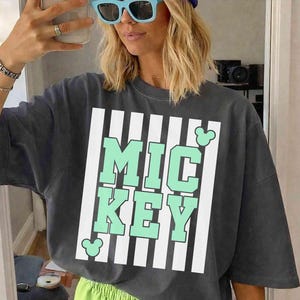 K&ouml;nnte beinhalten: Dunkelgraues T-Shirt mit dem Wort "MICKEY" in mintgr&uuml;nen Blockbuchstaben, umrahmt von wei&szlig;en Streifen. Das Shirt wird mit neongr&uuml;nen Shorts kombiniert. Ein l&auml;ssiges, bequemes Outfit.