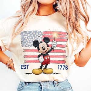 May include: Cream-colored t-shirt featuring a graphic of Mickey Mouse holding a sparkler in front of an American flag. The shirt has the text "EST 1776" in blue. The design is a patriotic theme.