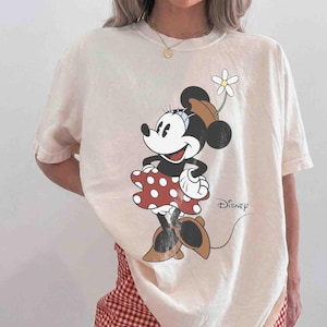 May include: Cream-colored t-shirt featuring a Minnie Mouse graphic. Minnie is depicted in a red polka-dot dress, brown shoes, and a hat with a flower. The Disney logo is printed on the shirt.