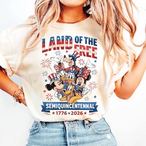 May include: Cream-colored t-shirt featuring a patriotic design with Mickey Mouse, Minnie Mouse, Donald Duck, Daisy Duck, Goofy, and Pluto. The text "LAND OF THE FREE" and "SEMIQUINCENTENNIAL 1776-2026" are displayed in red, white, and blue.