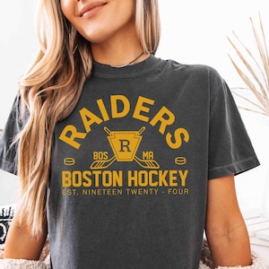 May include: Dark gray t-shirt with the text "RAIDERS BOSTON HOCKEY" in gold lettering. The design includes hockey sticks and a puck. The shirt also has the text "EST. NINETEEN TWENTY-FOUR".