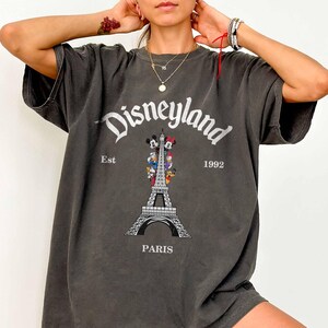May include: Dark gray oversized t-shirt featuring the Disneyland logo above a graphic of the Eiffel Tower with Mickey and Minnie Mouse. The text "Est 1992 PARIS" is also printed on the shirt.