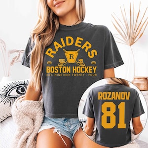 May include: Dark gray t-shirt with the text "RAIDERS BOSTON HOCKEY" in gold. The back of the shirt displays "ROZANOV 81" in gold. The shirt is a vintage style and features a hockey-themed design.