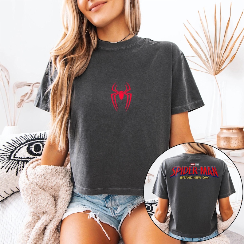 May include: Dark gray t-shirt featuring a red Spider-Man logo on the front. The back of the shirt displays the text "Spider-Man Brand New Day" in red and yellow. The shirt is a crew neck and has short sleeves.