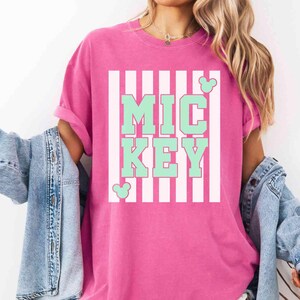 K&ouml;nnte beinhalten: Ein pinkfarbenes T-Shirt mit dem Wort "MICKEY" in mintgr&uuml;nen Blockbuchstaben, vor einem wei&szlig;-rosa gestreiften Hintergrund. Mickey Mouse-Silhouetten sind auf dem Shirt. Das Shirt ist Teil einer Casual-Wear-Kollektion.