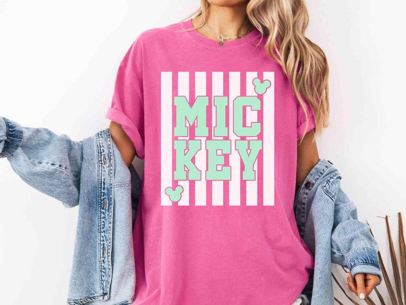 K&ouml;nnte beinhalten: Ein pinkfarbenes T-Shirt mit dem Wort "MICKEY" in mintgr&uuml;nen Blockbuchstaben, vor einem wei&szlig;-rosa gestreiften Hintergrund. Mickey Mouse-Silhouetten sind auf dem Shirt. Das Shirt ist Teil einer Casual-Wear-Kollektion.