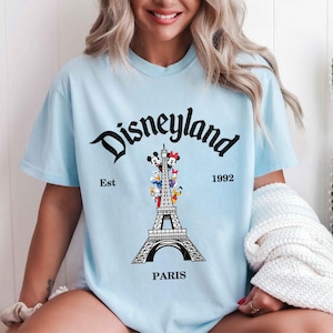 May include: Light blue t-shirt with a Disneyland Paris graphic. The design features the Eiffel Tower with Disney characters and the words "Disneyland Paris" and "Est 1992" in black script.