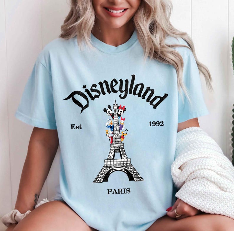 May include: Light blue t-shirt with a Disneyland Paris graphic. The design features the Eiffel Tower with Disney characters and the words "Disneyland Paris" and "Est 1992" in black script.