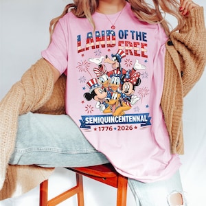 May include: Pink t-shirt with the text "LAND OF THE FREE" in red, white, and blue. Features cartoon characters Mickey Mouse, Minnie Mouse, Donald Duck, Goofy, and Pluto. The shirt also has the text "SEMIQUINCENTENNIAL 1776-2026".