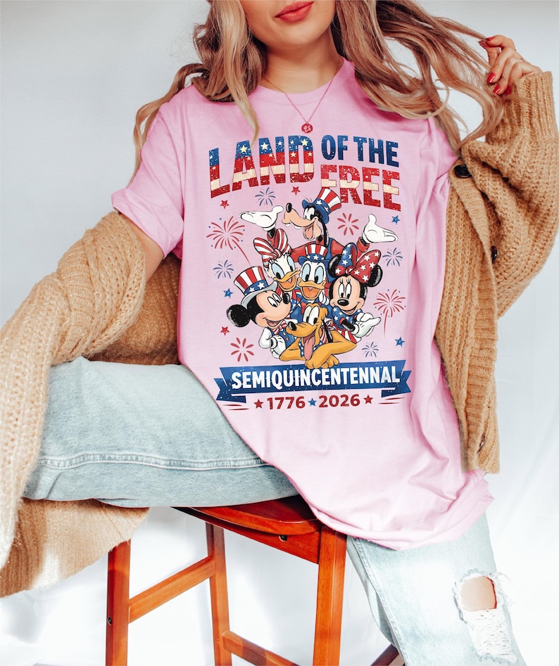 May include: Pink t-shirt with the text "LAND OF THE FREE" in red, white, and blue. Features cartoon characters Mickey Mouse, Minnie Mouse, Donald Duck, Goofy, and Pluto. The shirt also has the text "SEMIQUINCENTENNIAL 1776-2026".
