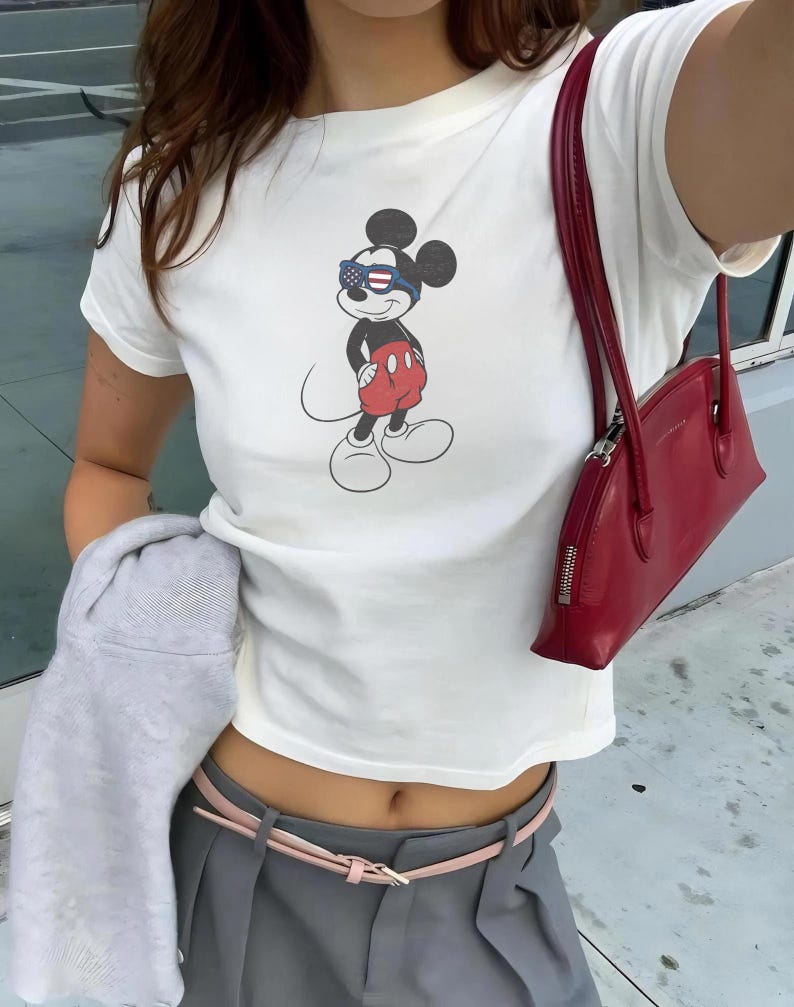 May include: A white short-sleeved t-shirt featuring a graphic of Mickey Mouse wearing sunglasses with an American flag pattern. The outfit includes grey trousers with a thin pink belt, a red shoulder bag, and a light grey garment held at the side.