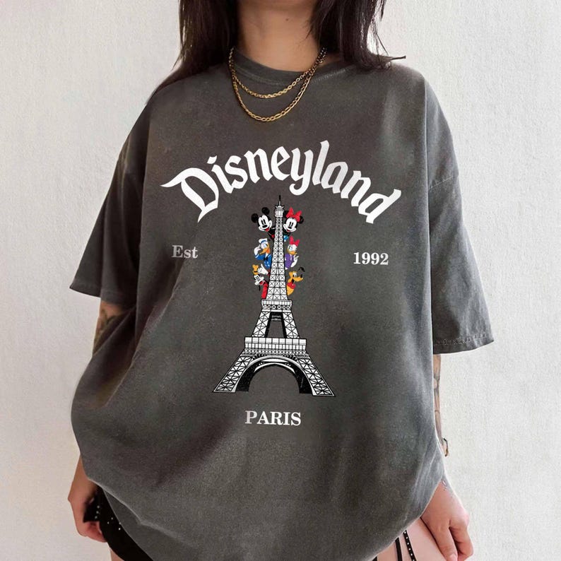 May include: Dark gray t-shirt featuring the word "Disneyland" in white script above an illustration of the Eiffel Tower with Disney characters. The text "Est 1992" and the word "Paris" are also printed on the shirt.