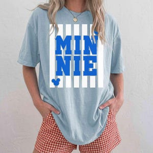 May include: Light blue t-shirt with the word "MINNIE" in bold blue letters, set against a white striped background. A small Mickey Mouse head is visible below the text. The shirt is a casual style.