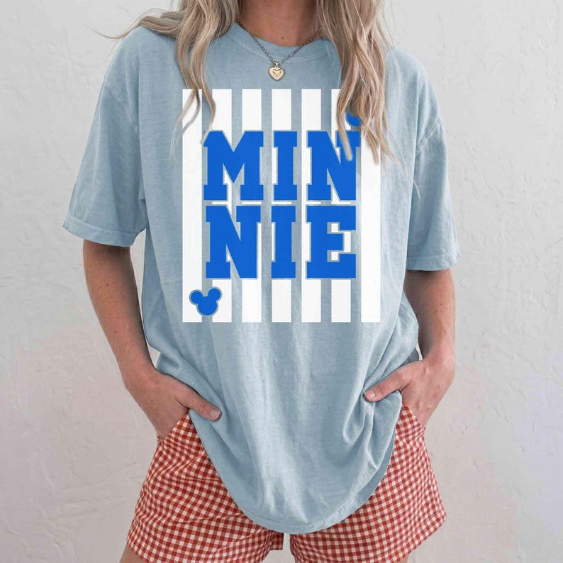 May include: Light blue t-shirt with the word "MINNIE" in bold blue letters, set against a white striped background. A small Mickey Mouse head is visible below the text. The shirt is a casual style.
