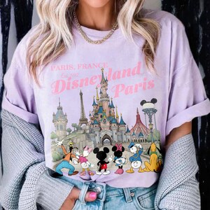 May include: A light purple t-shirt featuring a graphic of Disneyland Paris with Mickey Mouse, Minnie Mouse, Donald Duck, Goofy, and Pluto. The shirt has the text "Paris, France Disneyland Paris Est. 1992".