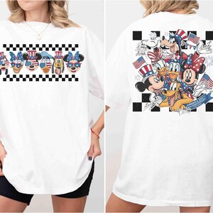 May include: White t-shirt featuring a graphic of Disney characters in patriotic attire. The front displays character faces with sunglasses, while the back showcases a group of characters with flags and fireworks. A black and white checkered pattern frames the designs.