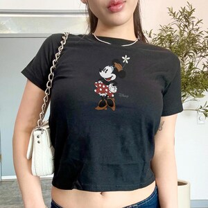 May include: A black short-sleeved t-shirt featuring a graphic of Minnie Mouse wearing a red and white polka dot skirt, brown shoes, and a hat with a white flower. The Disney logo is printed in small grey text below the character illustration.