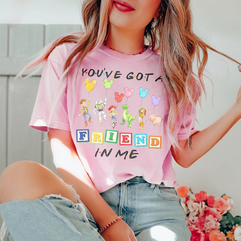 May include: A light pink short-sleeve t-shirt featuring a graphic of Toy Story characters holding Mickey Mouse-shaped balloons. The text reads YOU'VE GOT A FRIEND IN ME, with the word FRIEND displayed on colorful alphabet blocks.