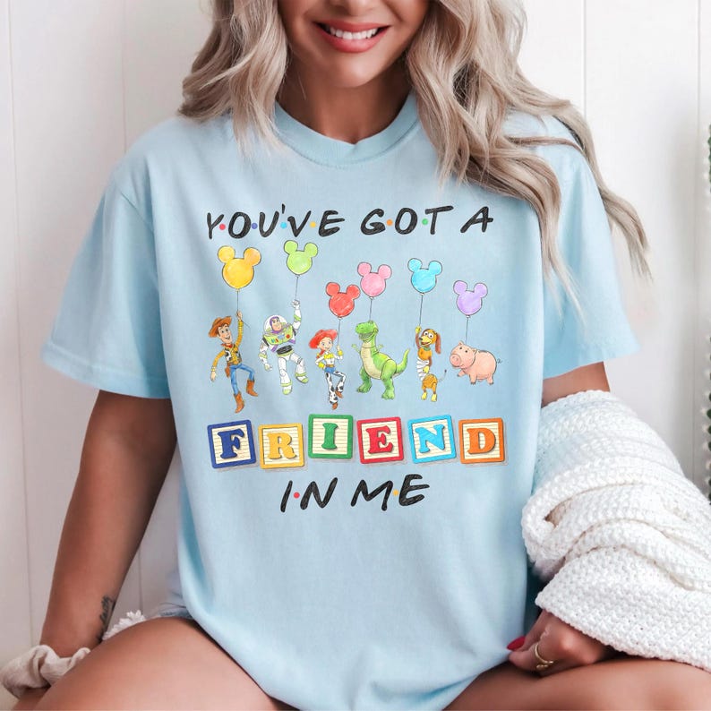 May include: A light blue t-shirt featuring a graphic of Toy Story characters holding Mickey Mouse-shaped balloons. The text reads YOU'VE GOT A FRIEND IN ME, with the word FRIEND spelled out in colorful alphabet blocks below the characters.