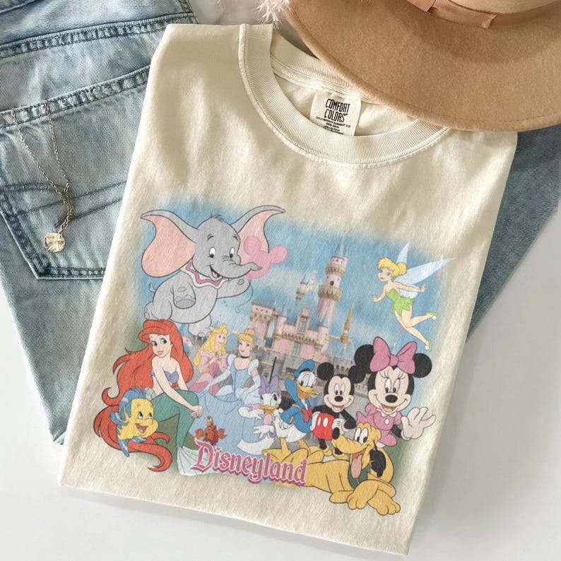 May include: Cream-colored t-shirt featuring a Disneyland graphic with cartoon characters like Dumbo, Ariel, Cinderella, Mickey and Minnie Mouse. The shirt is folded with a brown hat and blue jeans.