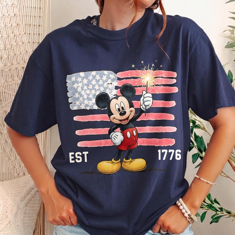 May include: Navy blue t-shirt featuring Mickey Mouse holding a sparkler in front of a stylized American flag. The shirt includes the text "EST 1776" in white, celebrating the founding of the United States.