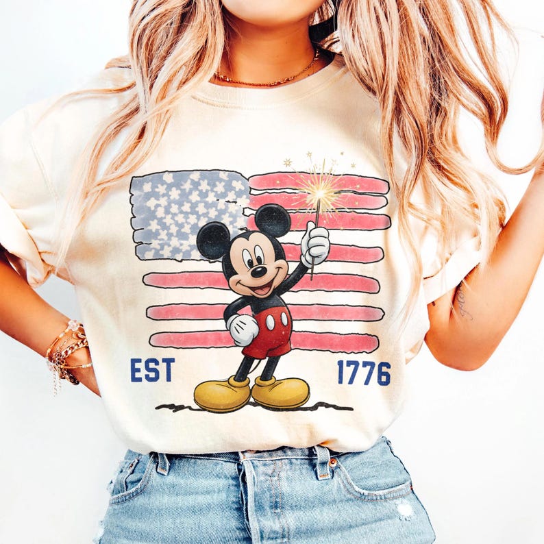 May include: Cream-colored t-shirt featuring a graphic of Mickey Mouse holding a sparkler in front of an American flag. The shirt has the text "EST 1776" in blue. The design is a patriotic theme.
