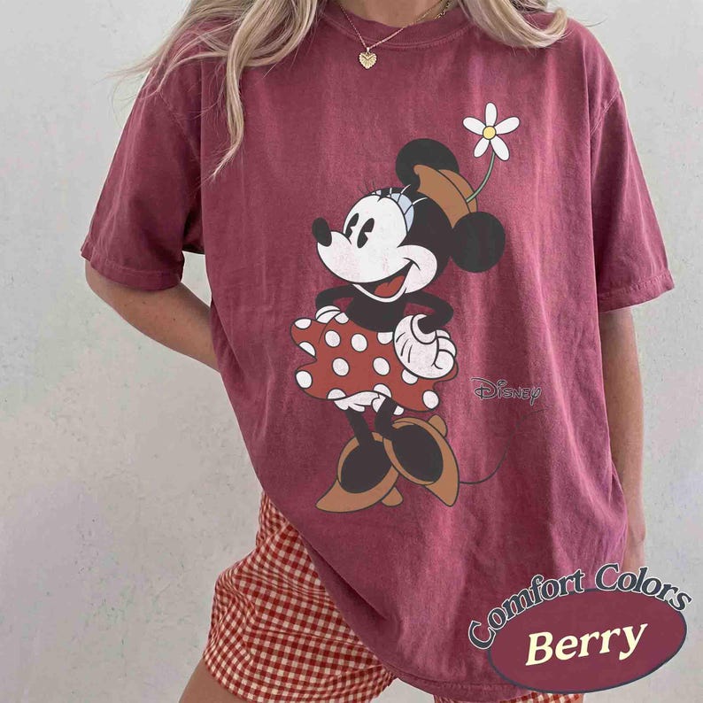 May include: Berry-colored t-shirt featuring a Minnie Mouse graphic. Minnie is wearing a red polka-dot dress and a brown hat with a white flower. The shirt has the words "Comfort Colors" and "Berry" printed on it, and the Disney logo.