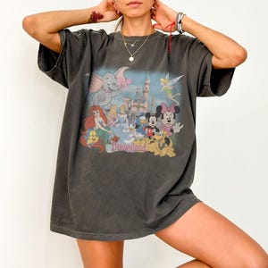 May include: Dark gray oversized t-shirt featuring a colorful graphic of Disney characters including Mickey and Minnie Mouse, Ariel, Dumbo, and Tinkerbell. The word "Disneyland" is printed below the characters. The shirt has short sleeves.